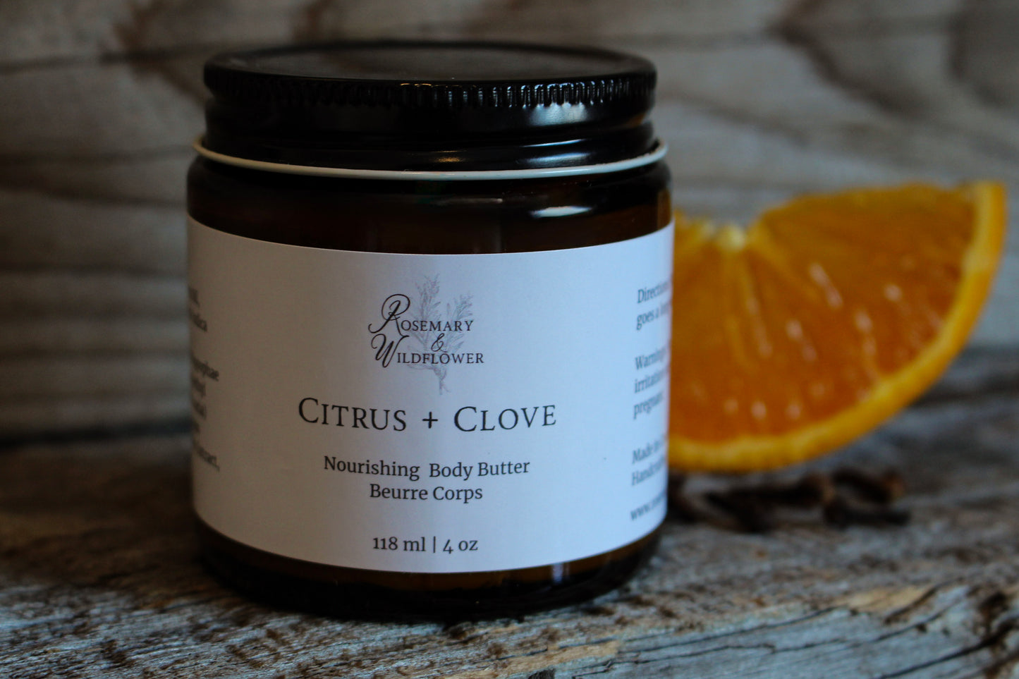 Jar of Citrus + Clove body butter with an orange slice and cloves on a wooden surface.