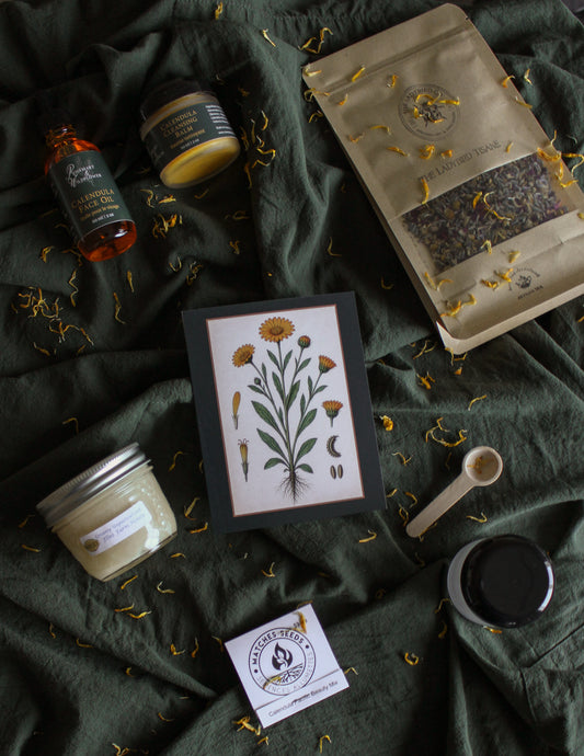 Calendula Box contents displayed on a green linen background with calendula petals strewn throughout.